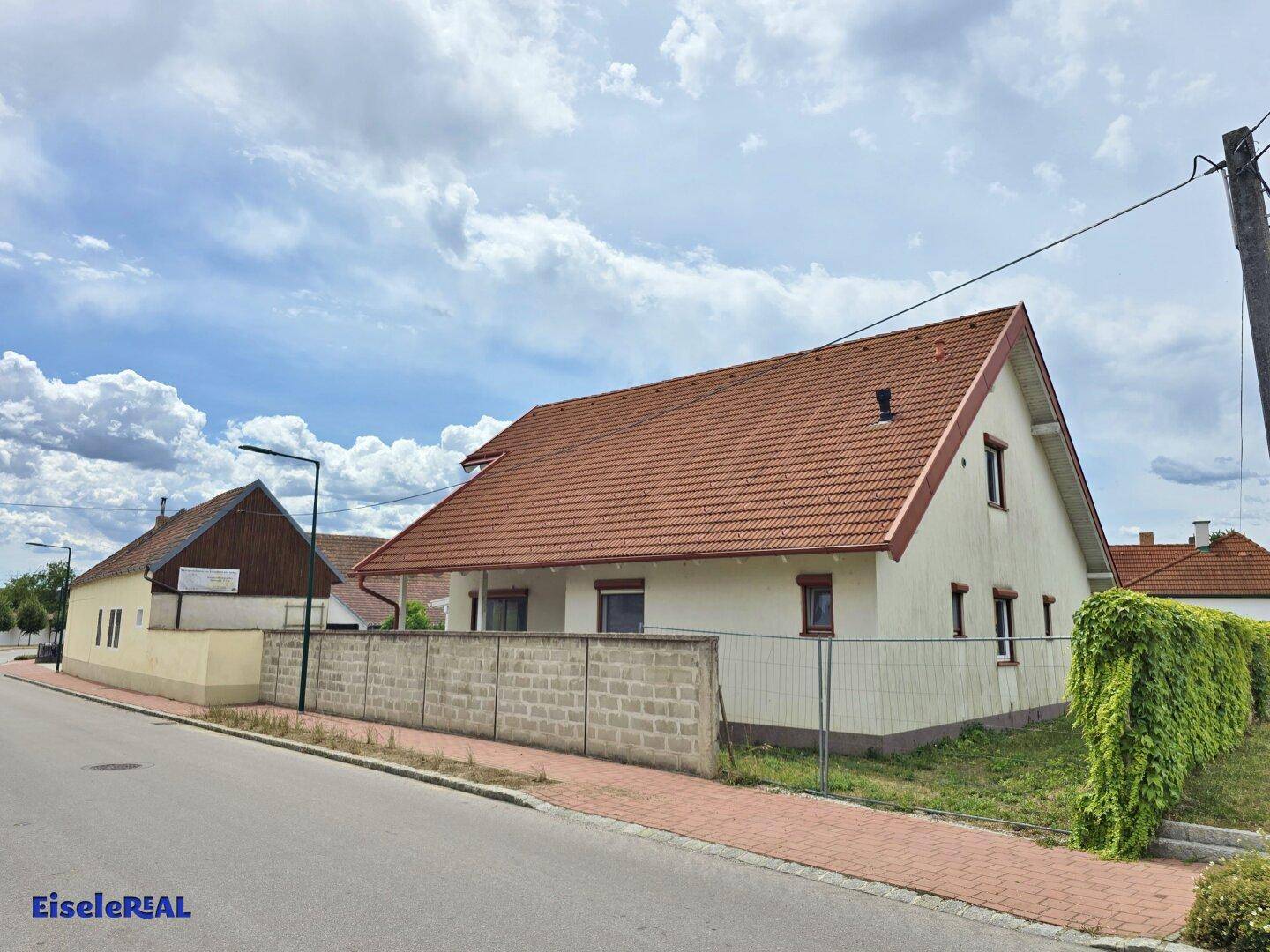 Wohnhaus mit rotem Ziegeldach, weißer Fassade und einer Betonmauer zur Straße hin.