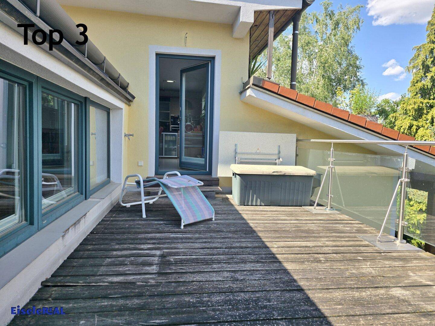 Geräumige Holzterrasse mit Sonnenliege und Zugang zum Innenbereich, umgeben von viel Grün.