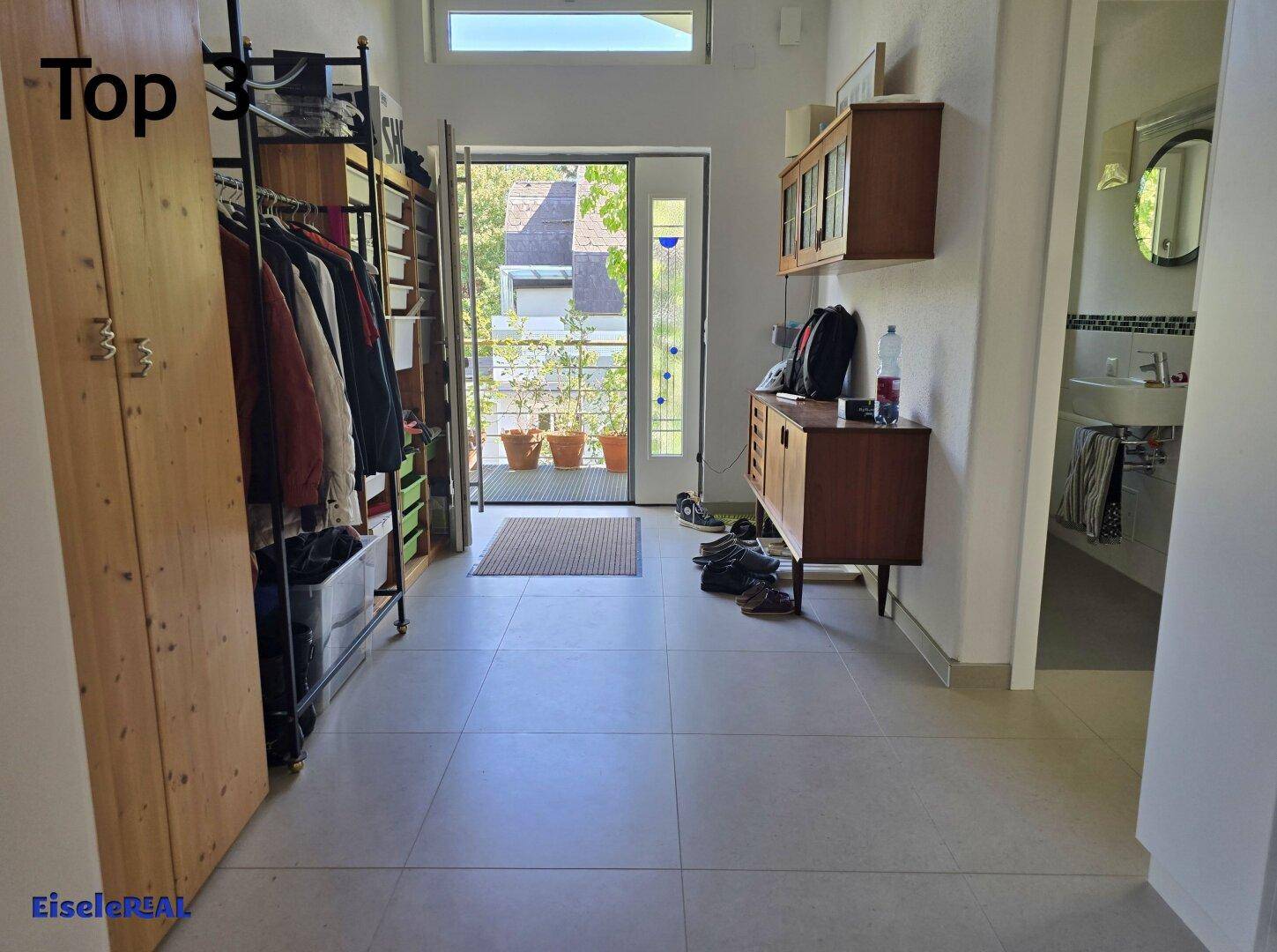 Heller Eingangsbereich mit Fliesenboden, Garderobe und Blick ins Grüne durch die Haustür.