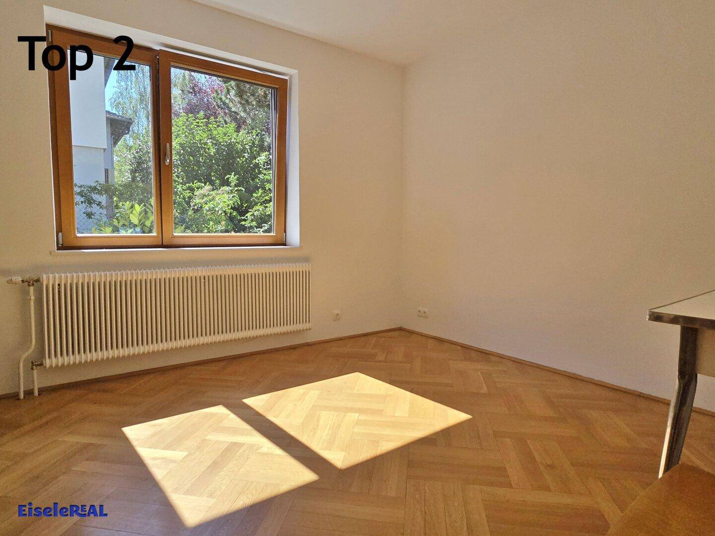 Helles Schlafzimmer mit Fischgrätparkett und großem Fenster, das viel Tageslicht hereinlässt.