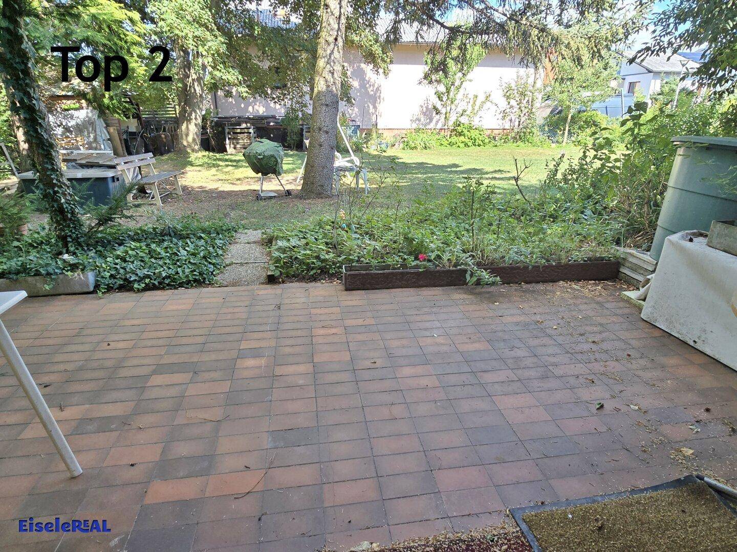 Gepflasterte Terrasse mit Gartenblick, umgeben von viel Grün und Bäumen.