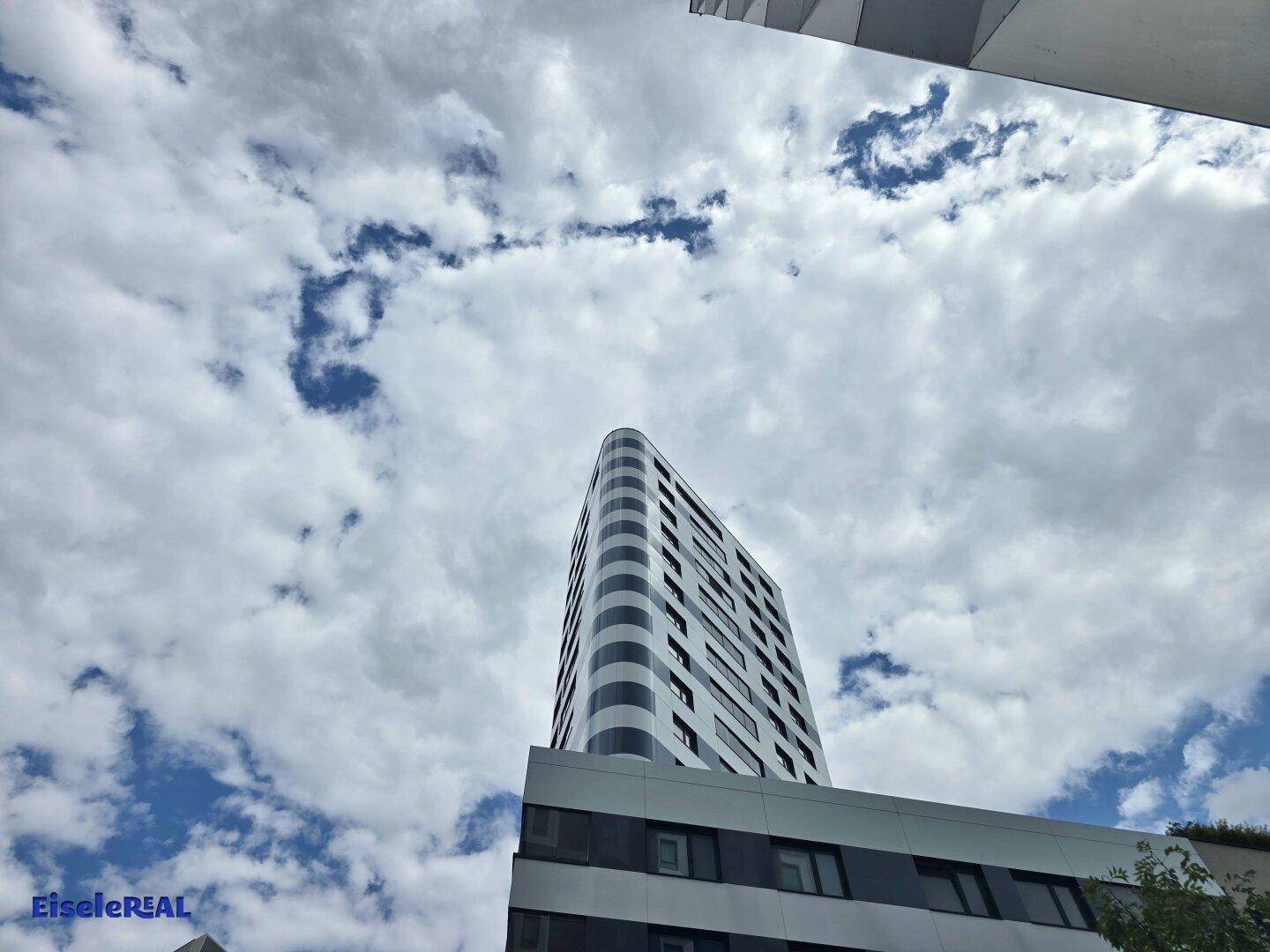 Hochhausfassade mit geschwungenen Linien und großen Fenstern unter einem teilweise bewölkten Himmel.