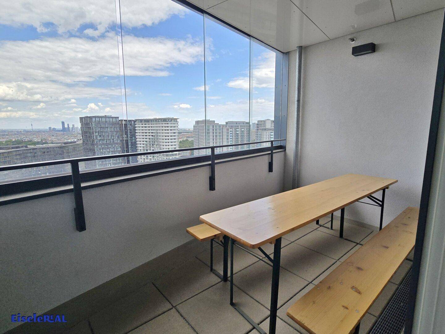 Verglaster Balkon mit Holztisch und Bänken, bietet einen weiten Blick über die Stadtlandschaft.