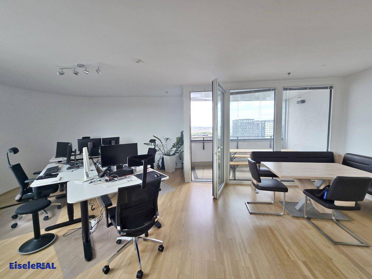 Offener Bürobereich mit Arbeitsplätzen, Essbereich und Zugang zu einem Balkon mit Stadtblick.