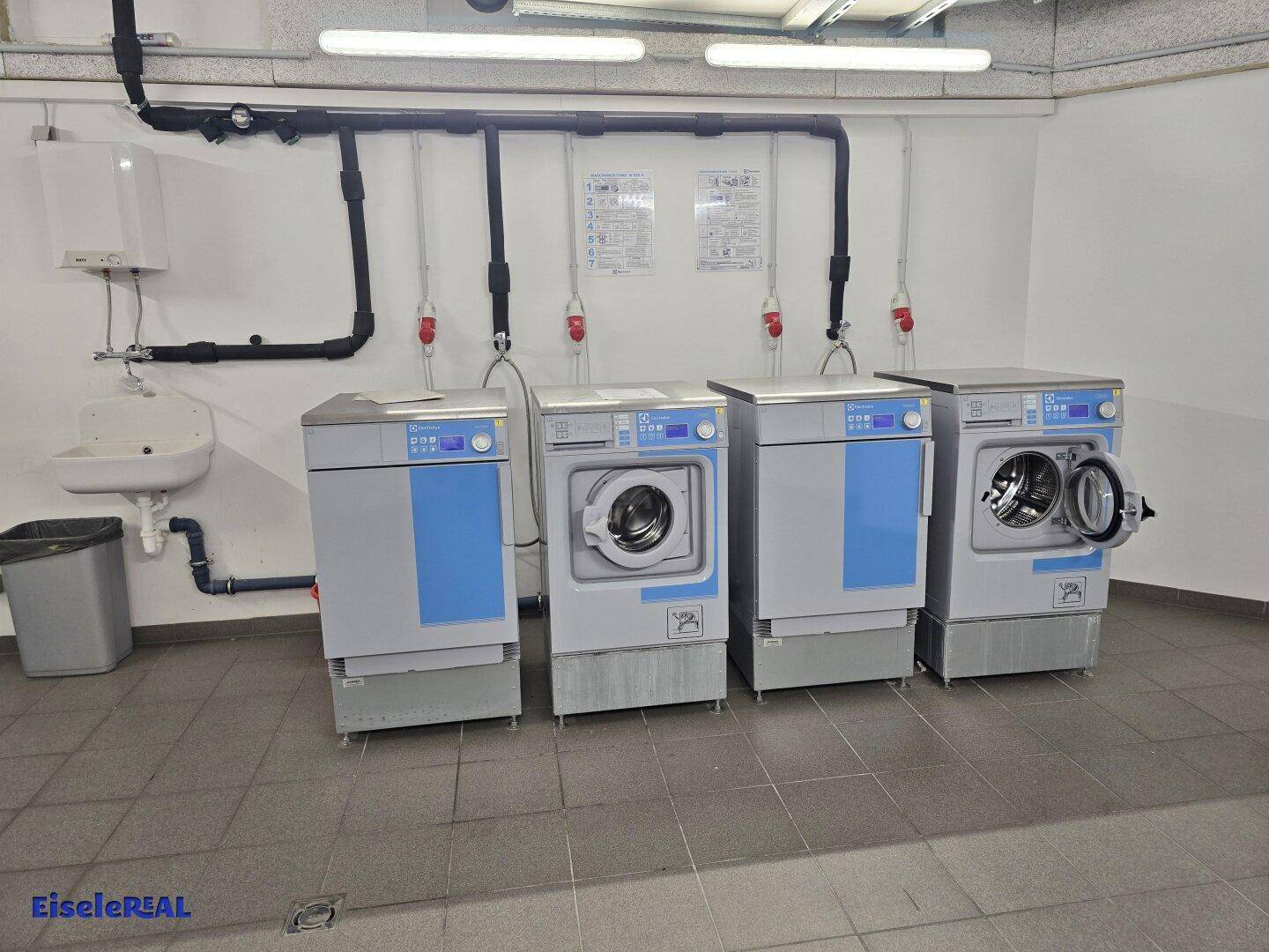 Gemeinschaftlicher Waschraum mit mehreren Waschmaschinen und einem Waschbecken, gefliester Boden.