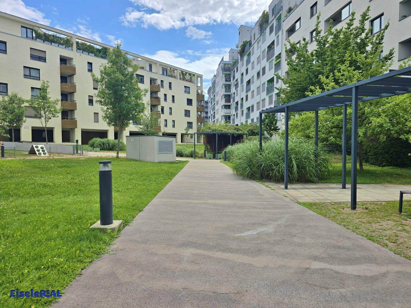 Gepflasterter Weg durch eine grüne Anlage mit Bäumen und modernen Wohngebäuden im Hintergrund.