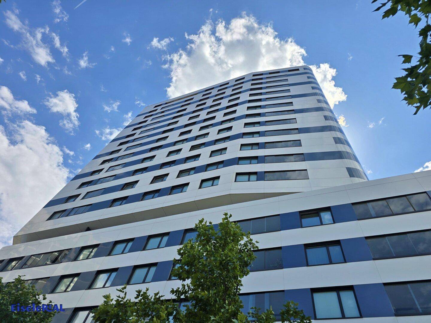 Ansicht eines modernen Hochhauses mit blauen und weißen Fassadenelementen unter bewölktem Himmel.