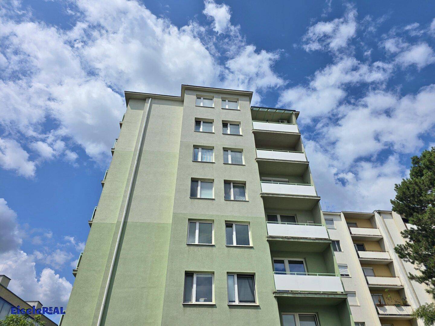 Ansicht eines modernen Wohnhauses mit hellgrüner Fassade und mehreren Balkonen unter blauem Himmel.
