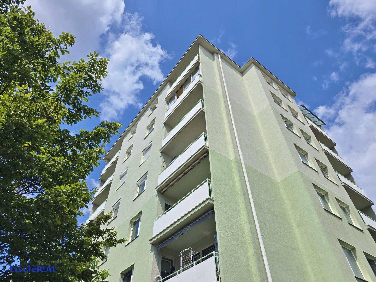 Fassade eines Wohngebäudes mit hellgrüner Farbgebung und Balkonen, umgeben von Bäumen.