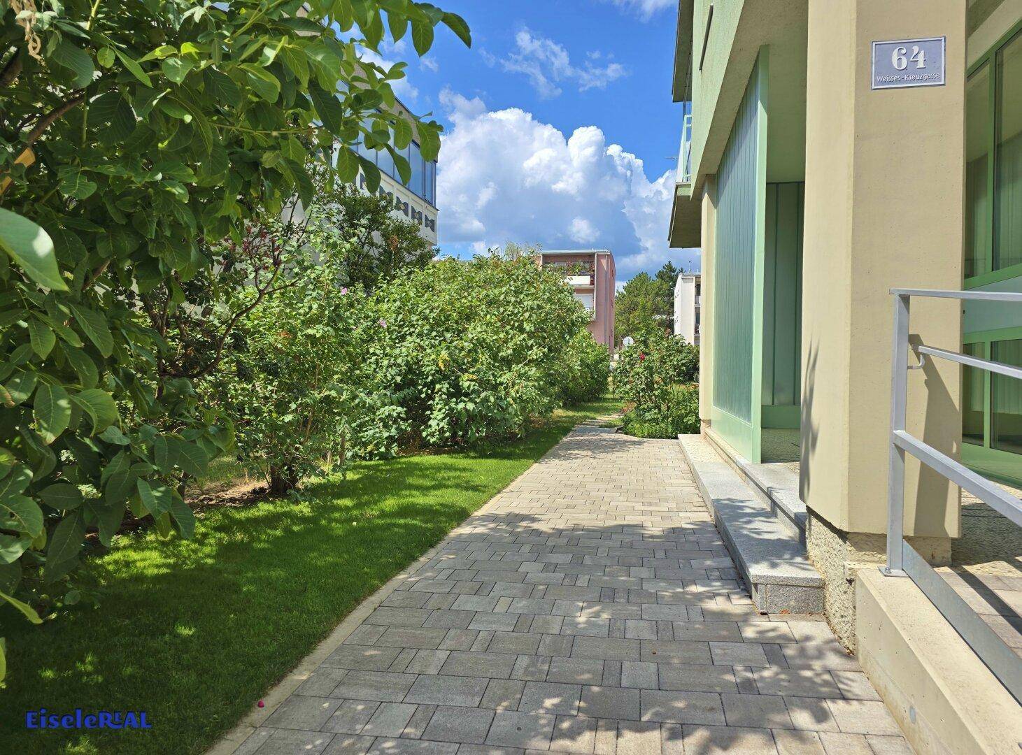 Gepflasterter Weg entlang eines Gebäudes mit grüner Rasenfläche und Sträuchern an einem sonnigen Tag.