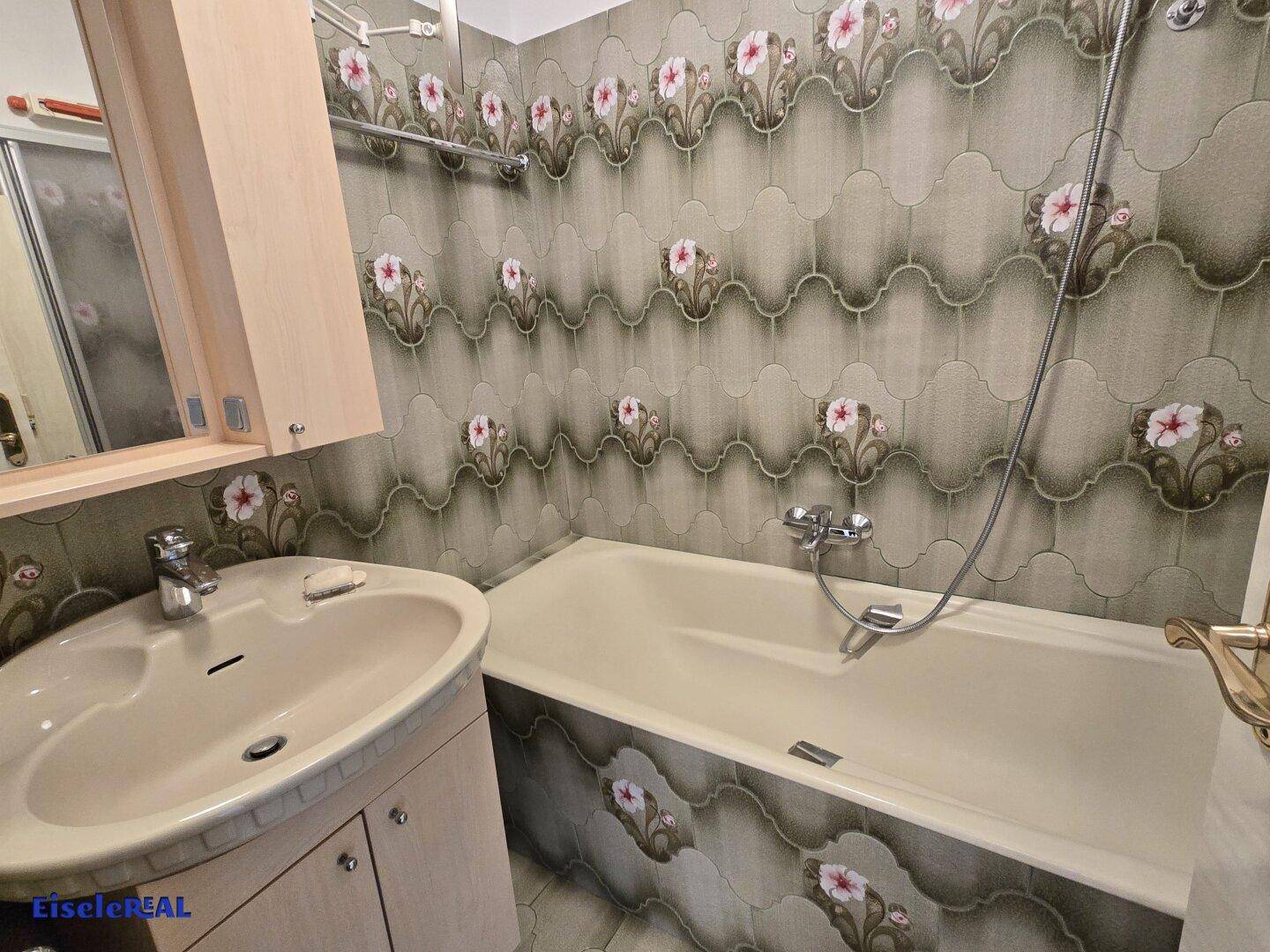 Badezimmer mit Badewanne, Waschbecken und floral gemusterten Fliesen.