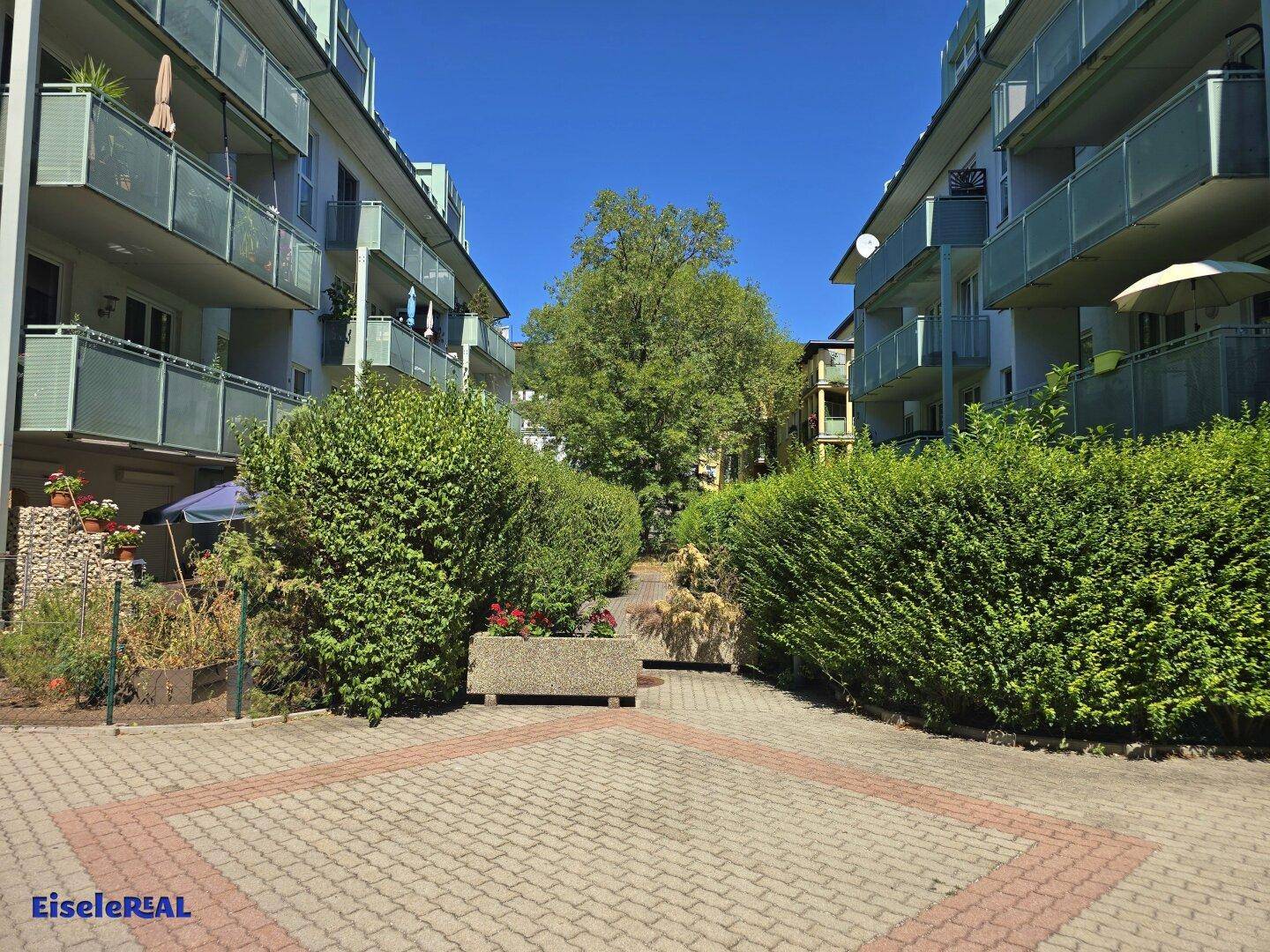 Gepflasterter Weg führt durch einen grünen Gartenbereich zwischen zwei Wohngebäuden an einem sonnigen Tag.