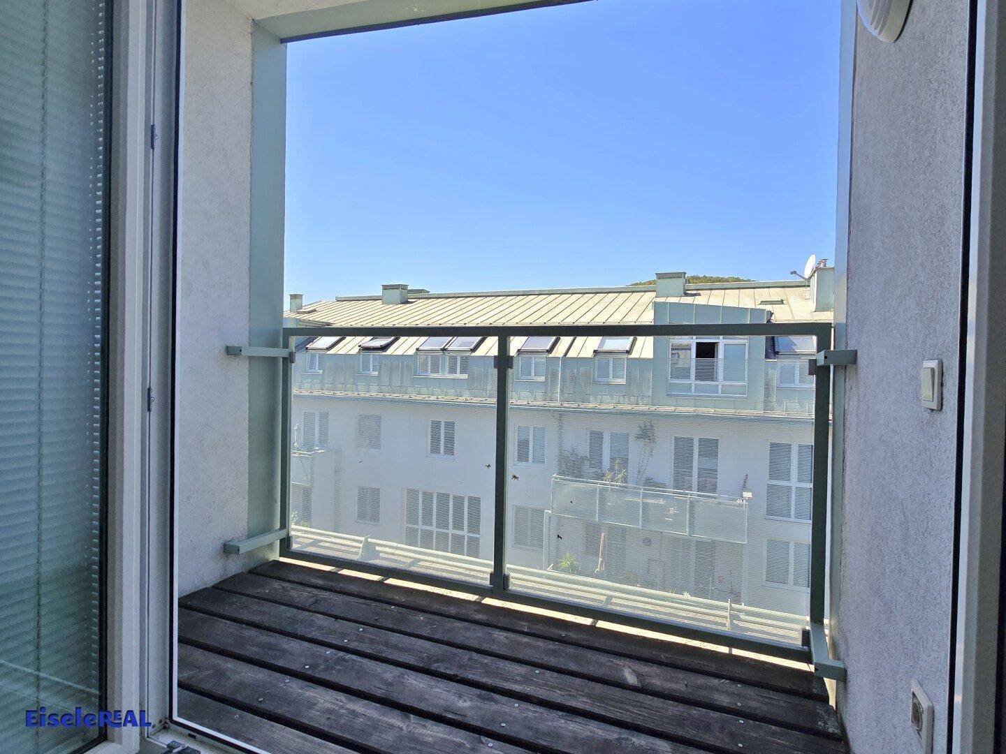 Kleiner Balkon mit Holzboden und Glasgeländer, bietet einen Blick auf die umliegenden Gebäude.