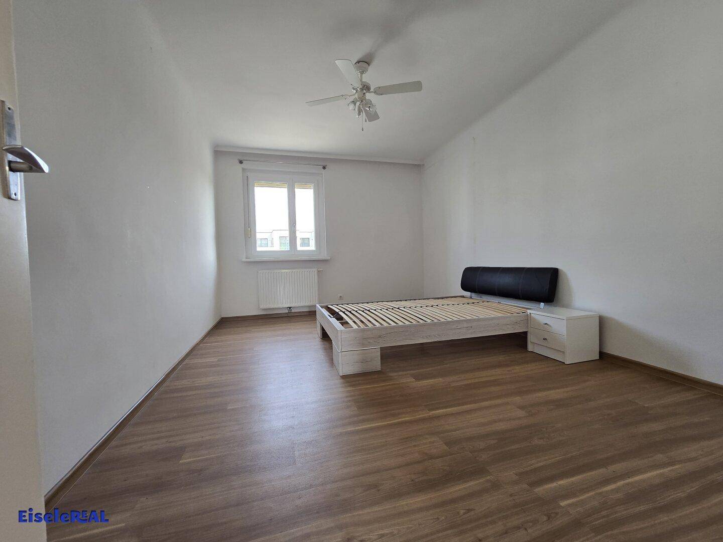 Schlafzimmer mit Laminatboden, Fenster und einem einfachen Bettgestell.