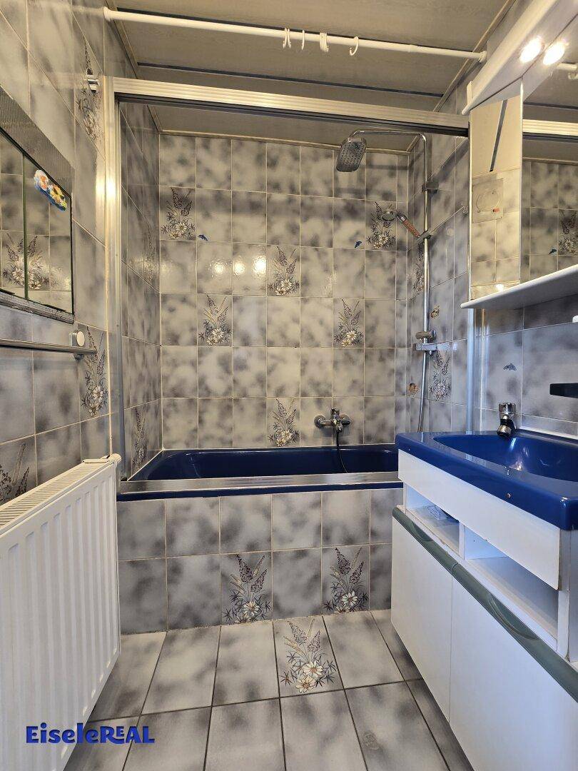 Badezimmer mit blauer Badewanne, Waschbecken und gemusterten Fliesen im 80er-Jahre-Stil.