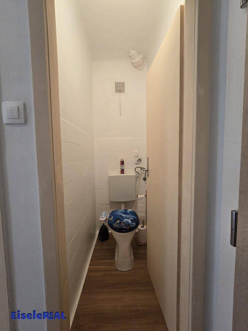 Separate Toilette mit WC, Fliesen an der Wand und Lüftungsgitter.