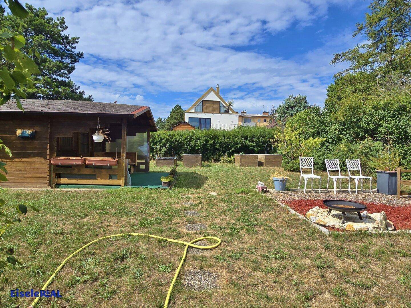 Garten mit einem charmanten Holzhaus, einer Feuerstelle und Blick auf ein modernes Gebäude.