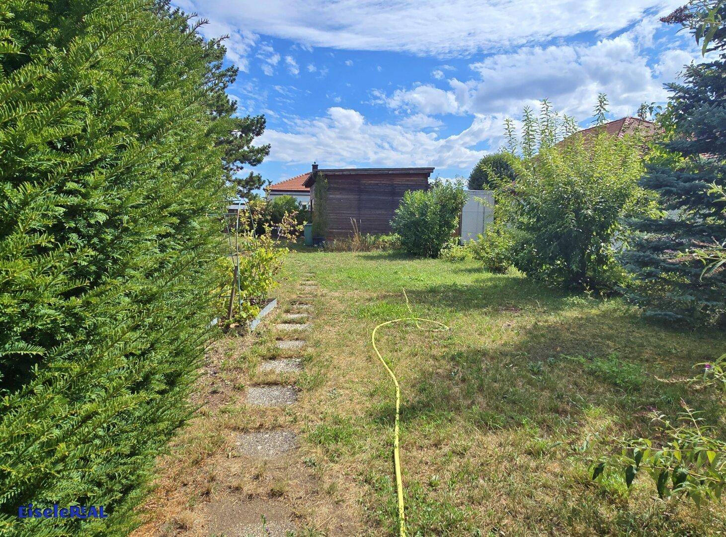 Gartenweg führt durch eine grüne Landschaft mit Bäumen und Sträuchern unter blauem Himmel.