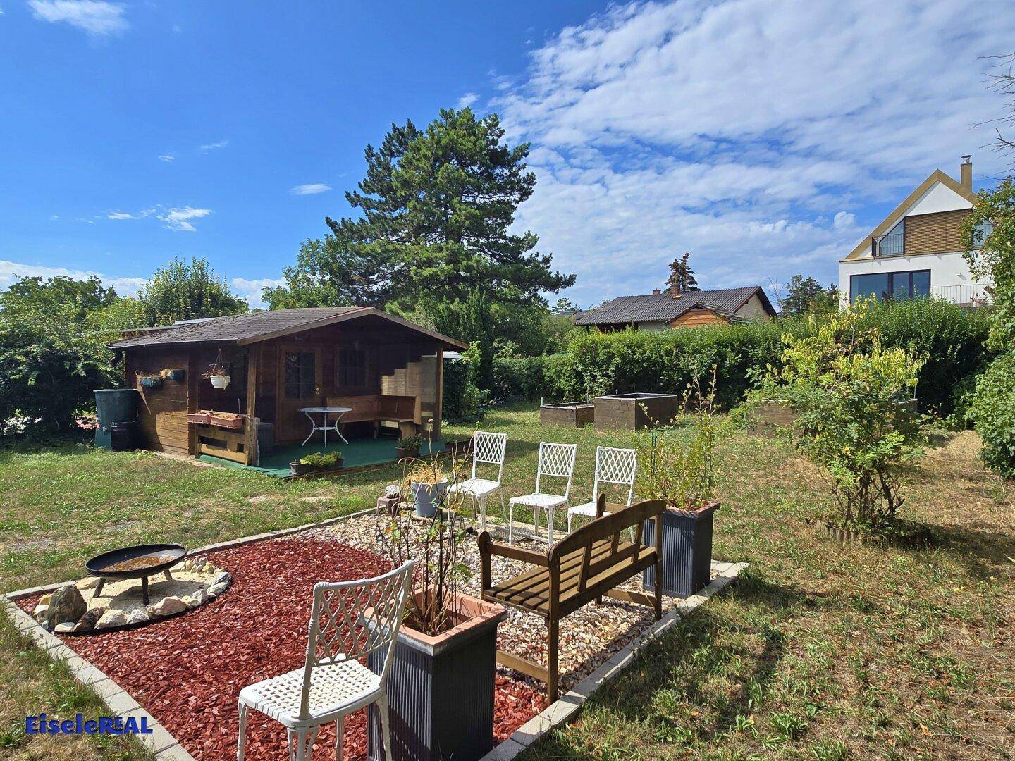Weitläufiger Garten mit Holzhaus, Feuerstelle und Sitzbereich, umgeben von Bäumen und Sträuchern.