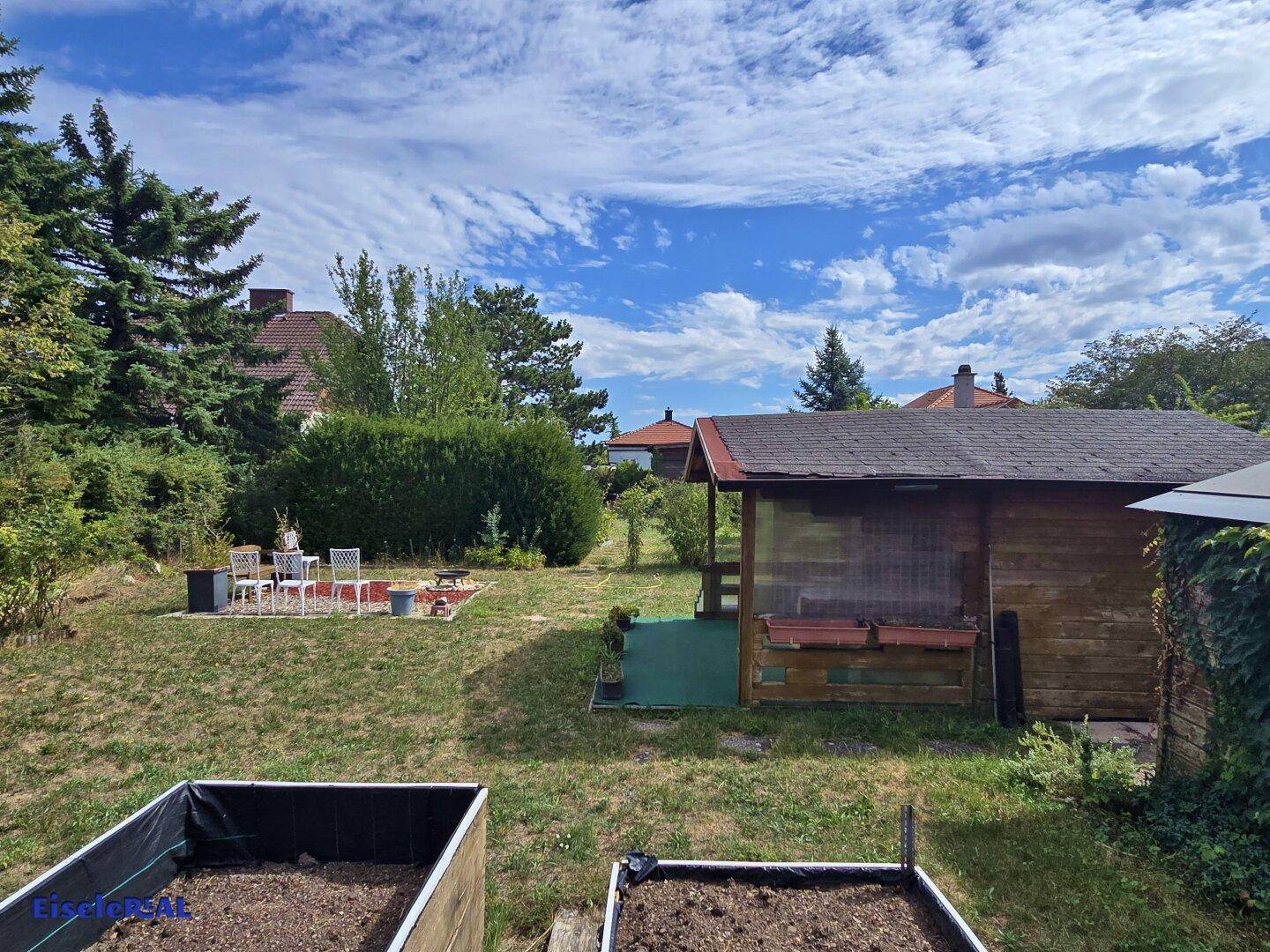 Garten mit Hochbeeten, einem Holzhaus und einer Feuerstelle, umgeben von üppigem Grün.