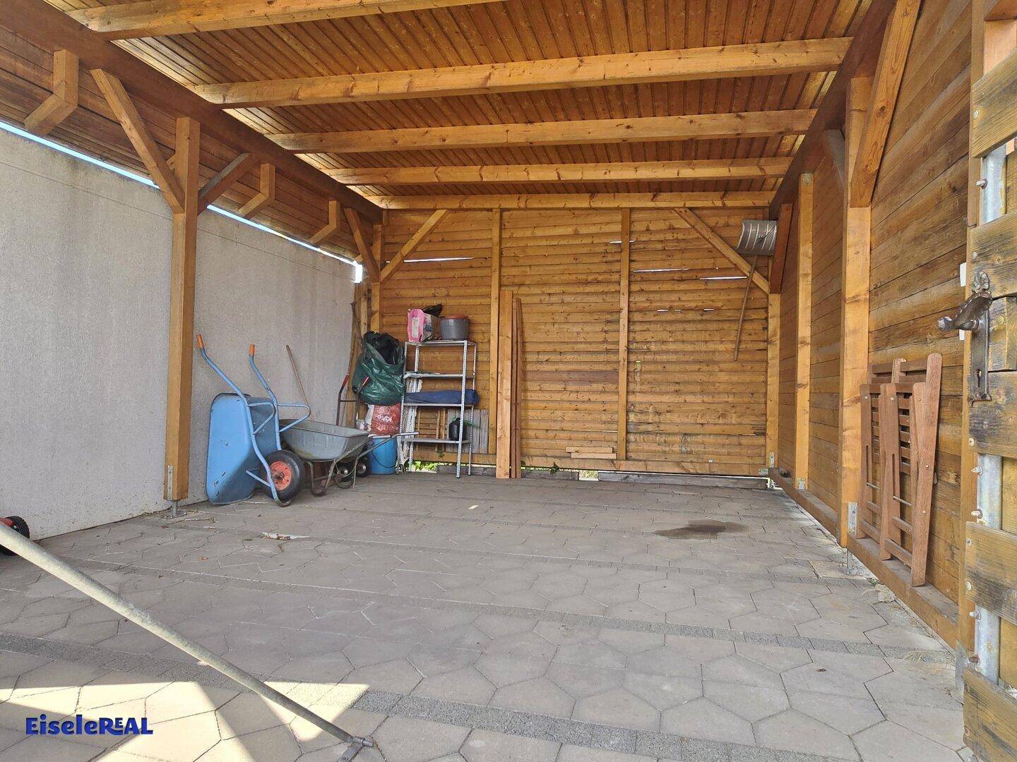 Überdachter Stellplatz oder Carport mit Holzkonstruktion und gepflastertem Boden.