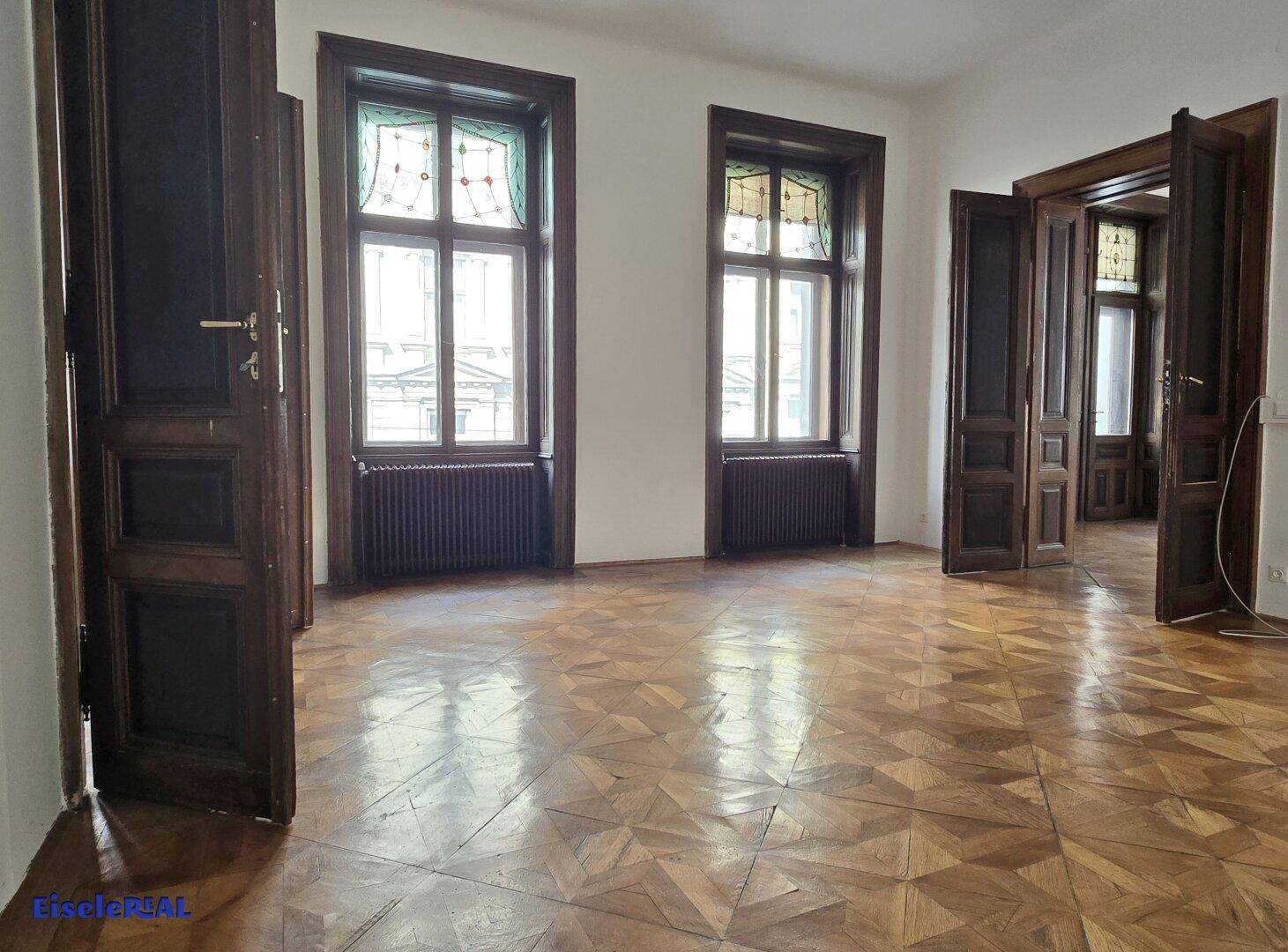 Helles Zimmer mit Parkettboden, zwei Fenstern mit Buntglas und dunklen Holztüren.