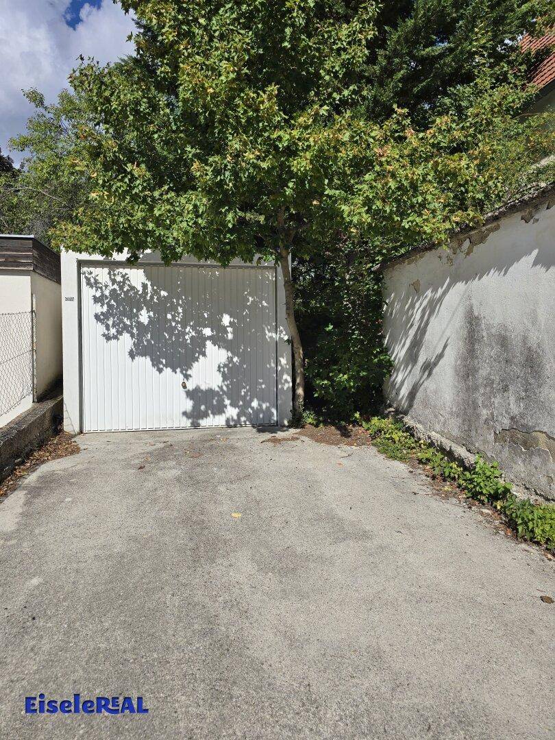 Einzelgarage mit weißem Tor und Zufahrt, umgeben von Bäumen und einer alten Mauer.
