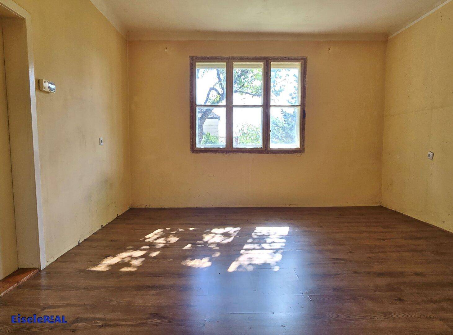 Geräumiger Raum mit gelben Wänden, großem Fenster und dunklem Laminatboden, lichtdurchflutet.