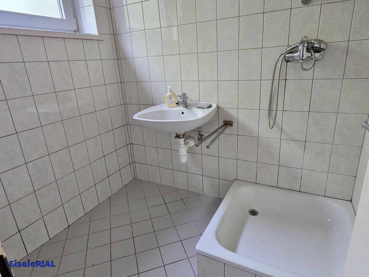 Badezimmer mit weißen Fliesen, Waschbecken und einer kleinen Badewanne mit Duscharmatur.