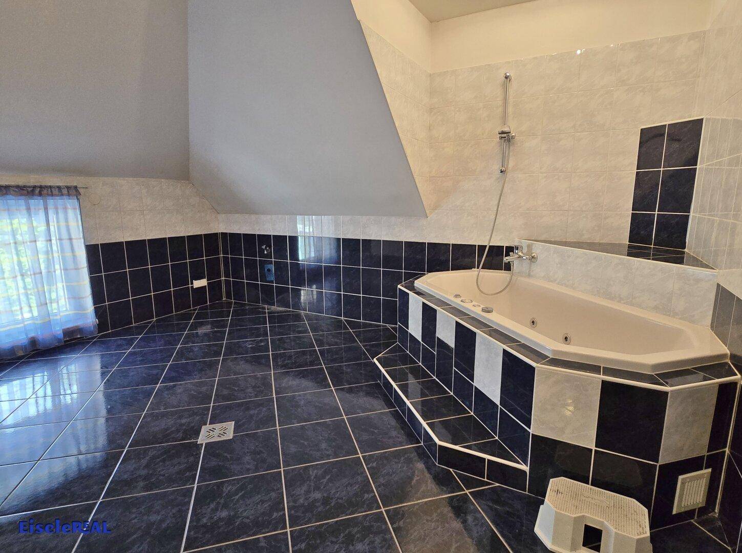 Geräumiges Badezimmer mit Eckbadewanne, dunklen Fliesen und schräger Decke.