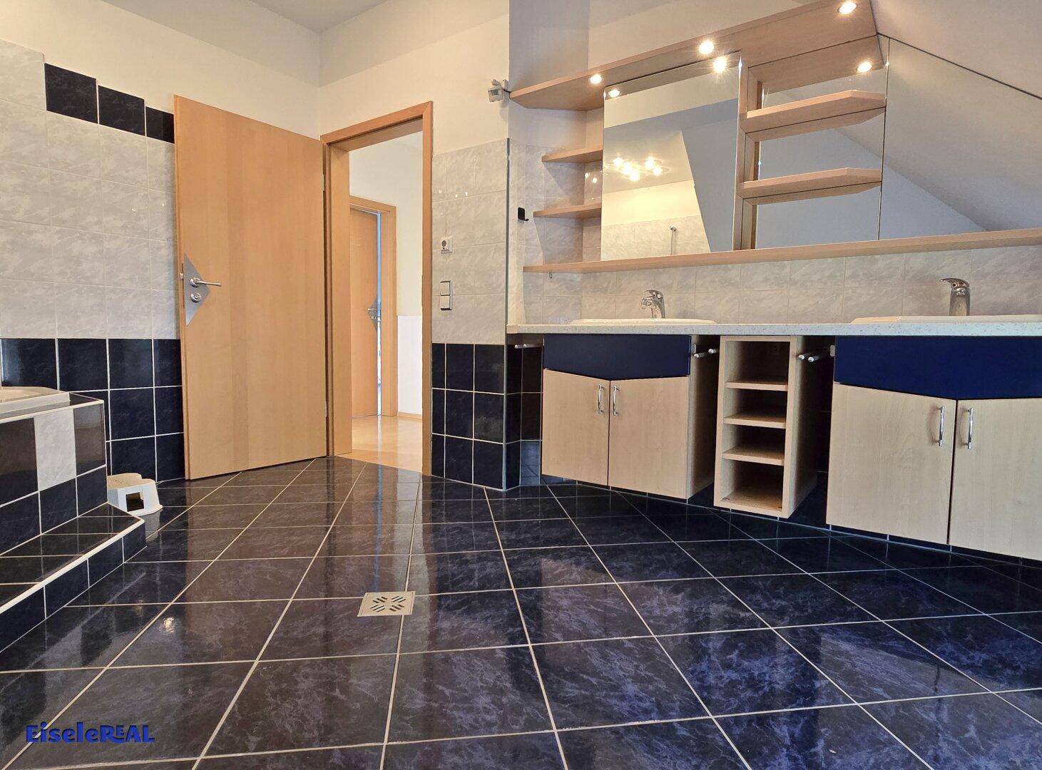 Badezimmer mit Doppelwaschbecken, Spiegelschrank und dunklen Fliesen.