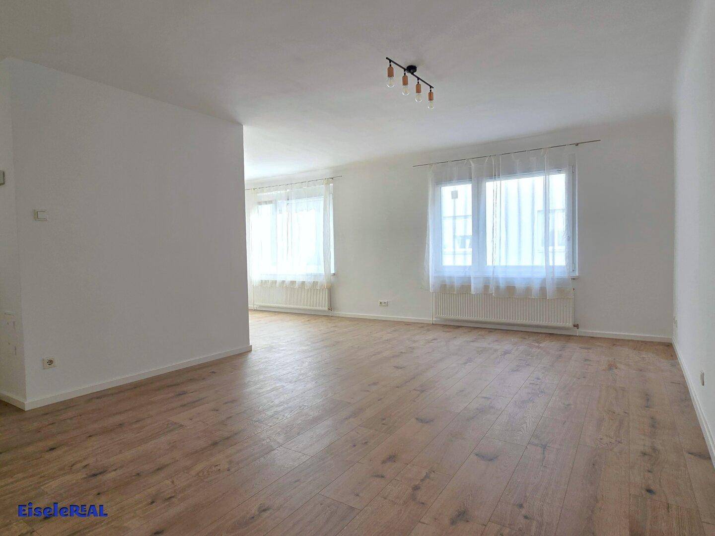Geräumiger, heller Raum mit zwei großen Fenstern und hellem Laminatboden, ideal als Wohnbereich.