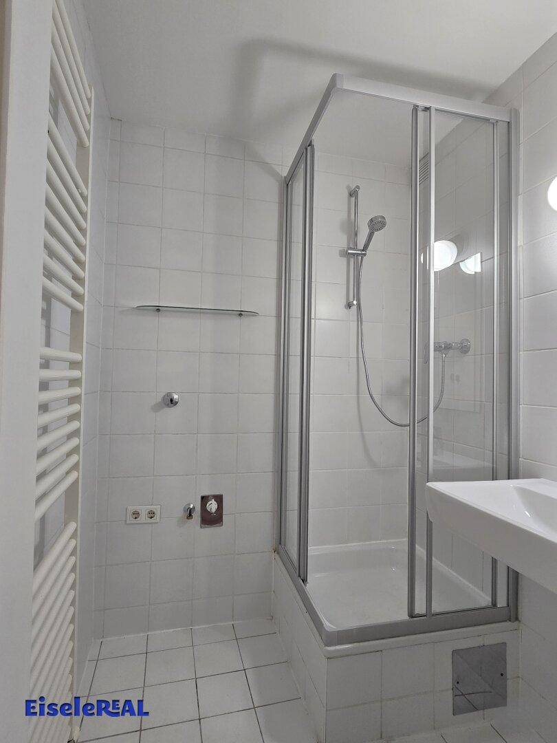 Helles Badezimmer mit weißen Fliesen, einer Duschkabine, Einzelwaschbecken und Handtuchheizkörper.