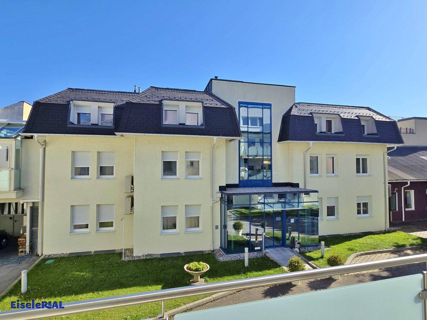 Rückansicht des Gebäudes mit Mansarddach und einem modernen Glasanbau im Eingangsbereich, umgeben von Grünflächen.