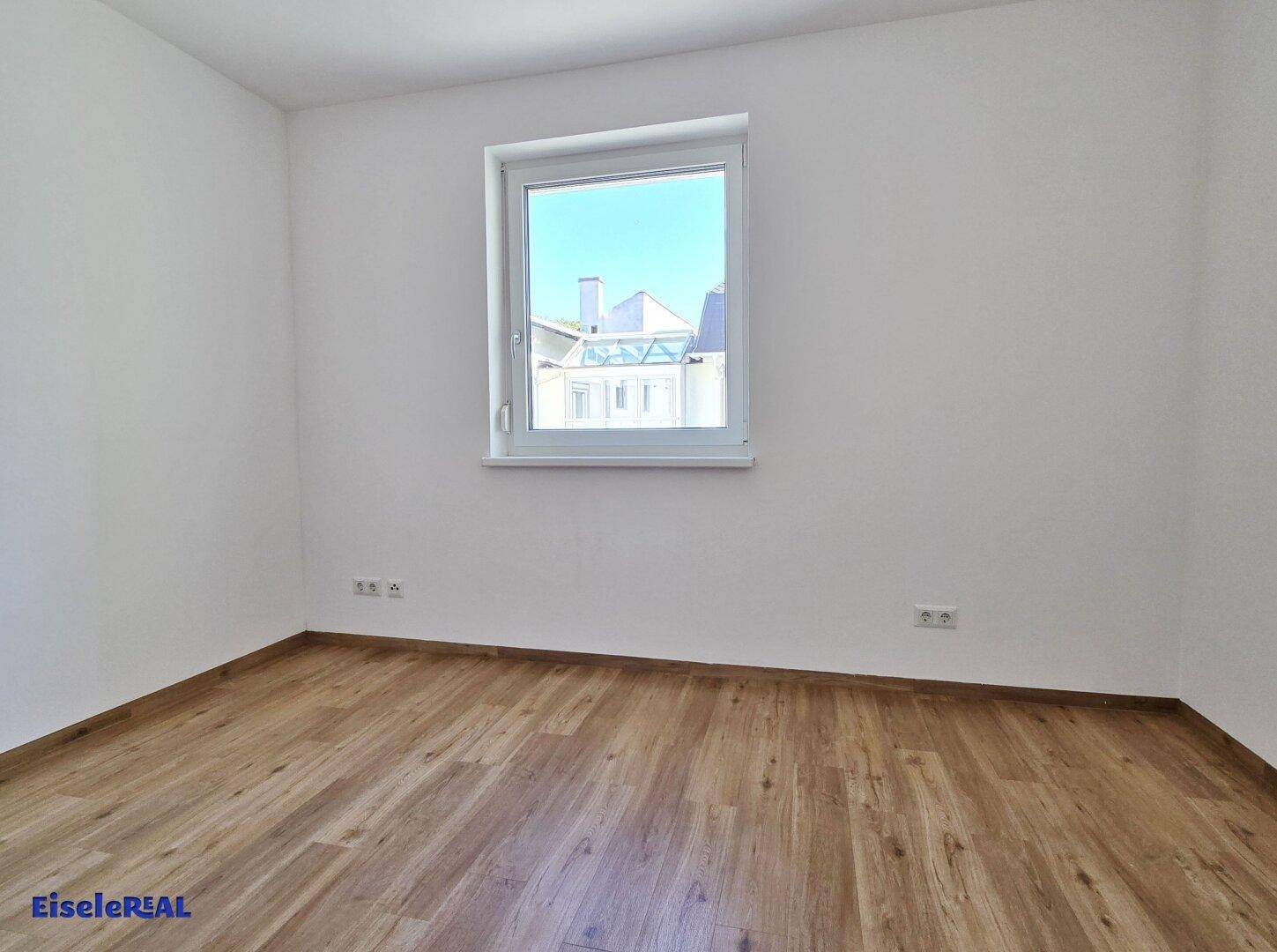 Helles Schlafzimmer mit Laminatboden und großem Fenster, das viel Tageslicht hereinlässt.