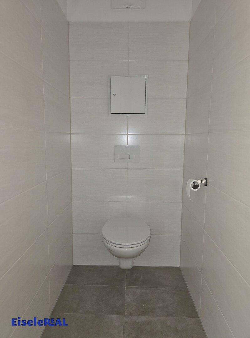 Modernes separates WC mit wandhängender Toilette und hellgrauen Fliesen an Wänden und Boden.