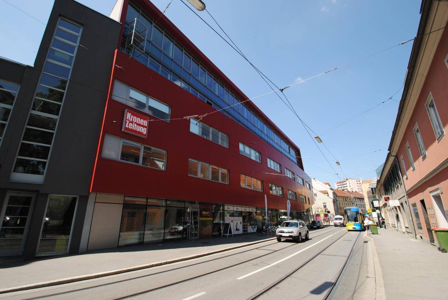 Modernes Geschäftsgebäude mit roter Fassade an einer belebten Straße mit Straßenbahn und Autos.