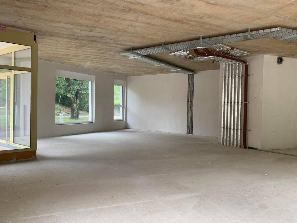 Unfertiger Innenraum mit freiliegender Betondecke, sichtbaren Installationen und großen Fenstern.
