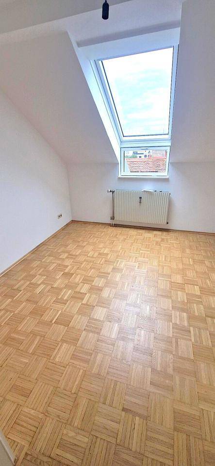 Helles Zimmer im Dachgeschoss mit Parkettboden, schrägen Wänden und einem großen Dachfenster.