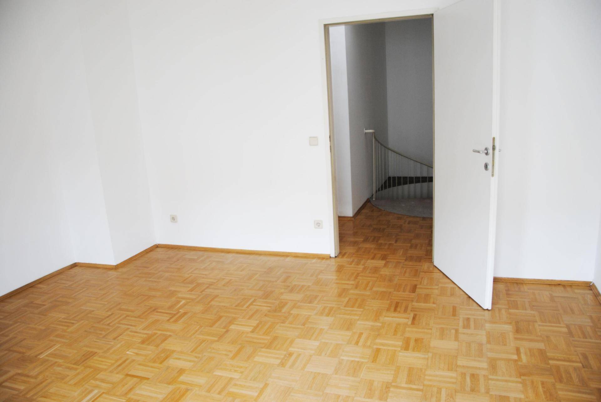 Leeres Zimmer mit Parkettboden und Zugang zu einer Wendeltreppe im Hintergrund.