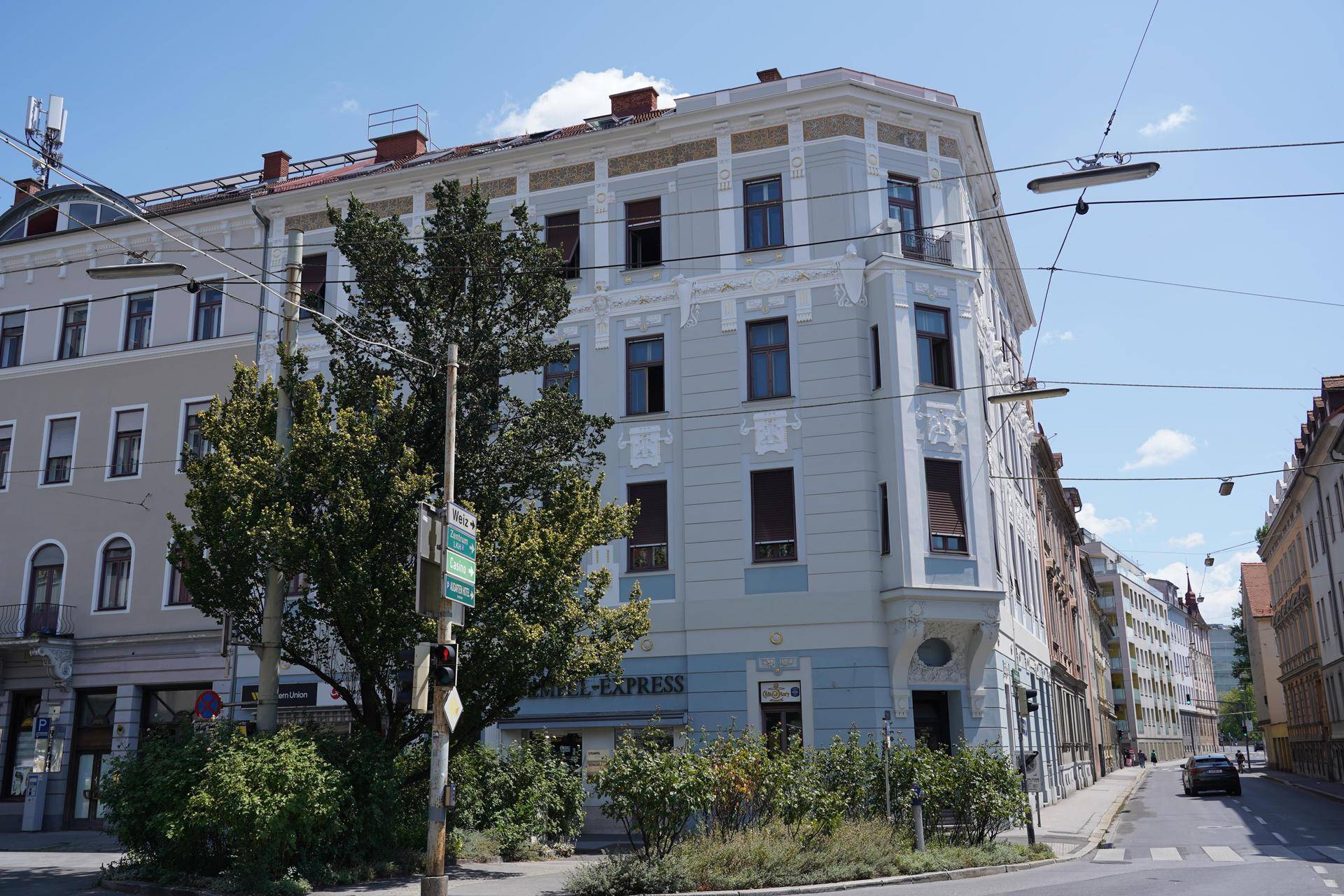 Historisches Eckgebäude mit blauer und weißer Fassade, umgeben von Bäumen und einer Straße.