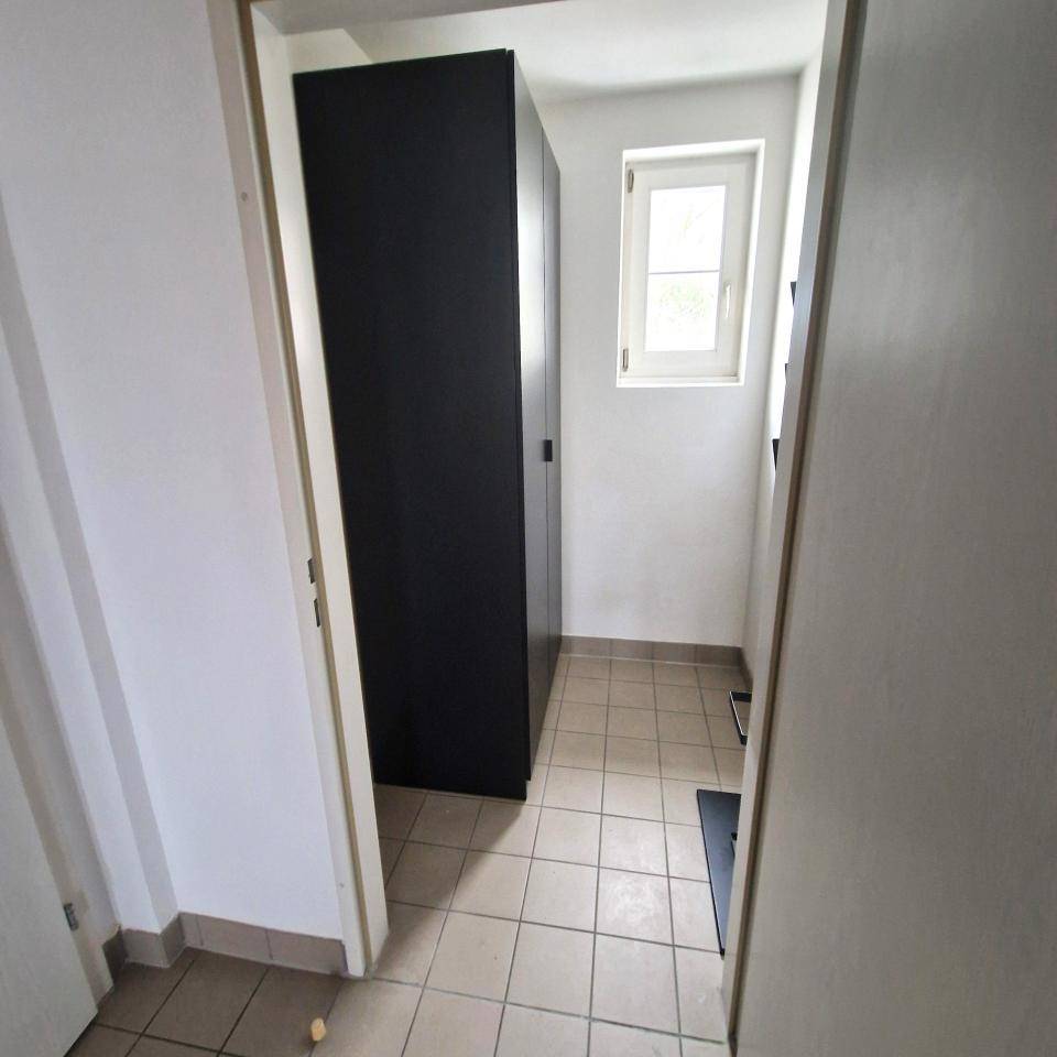 Heller Flur mit Fenster, Fliesenboden und einem schwarzen Schrank, der zusätzlichen Stauraum bietet.