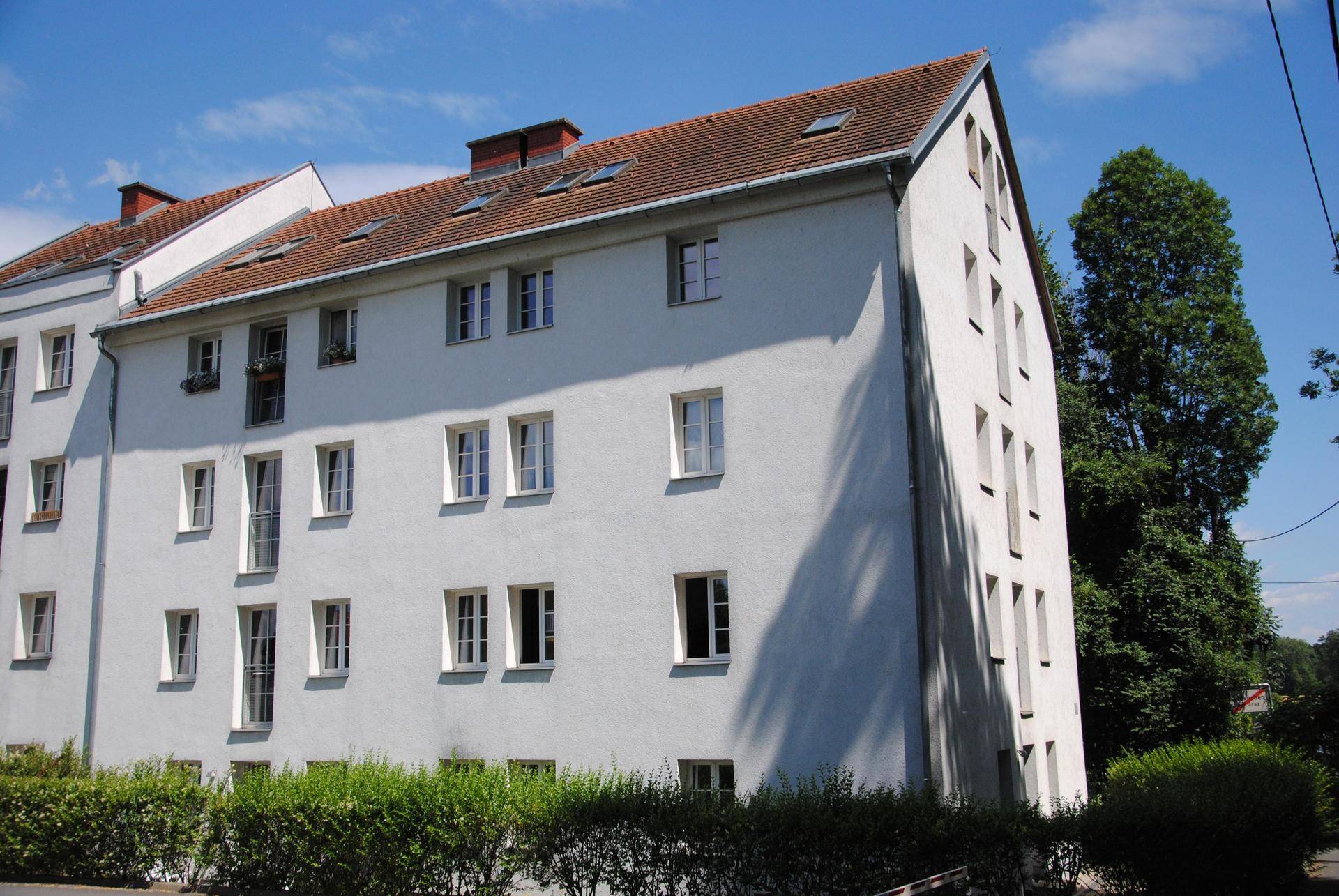 Mehrfamilienhaus mit weißer Fassade, Satteldach und vielen Fenstern, umgeben von Grünflächen.