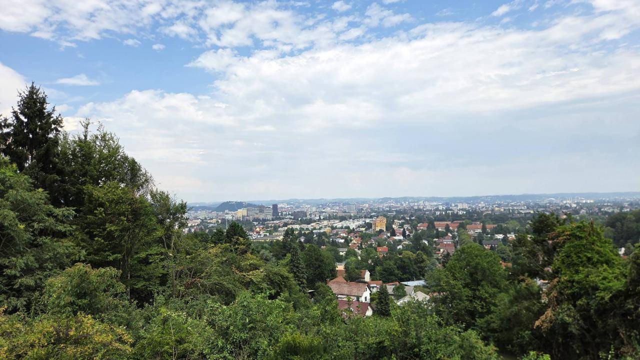 Weitläufiger Blick über eine grüne Hügellandschaft und die darunter liegende Stadt unter blauem Himmel.