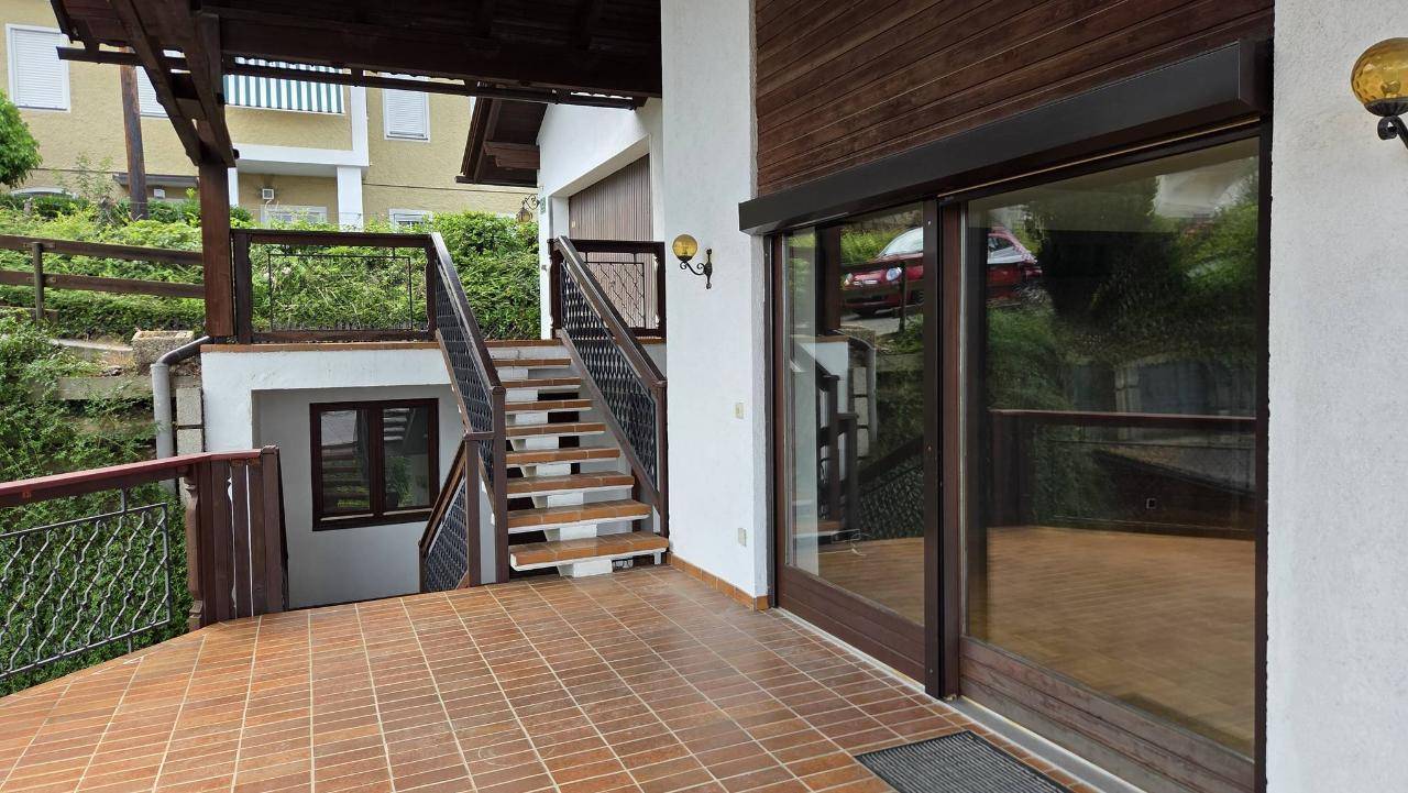Überdachte Terrasse mit Fliesenboden und Zugang zu einer Außentreppe, umgeben von Grün.