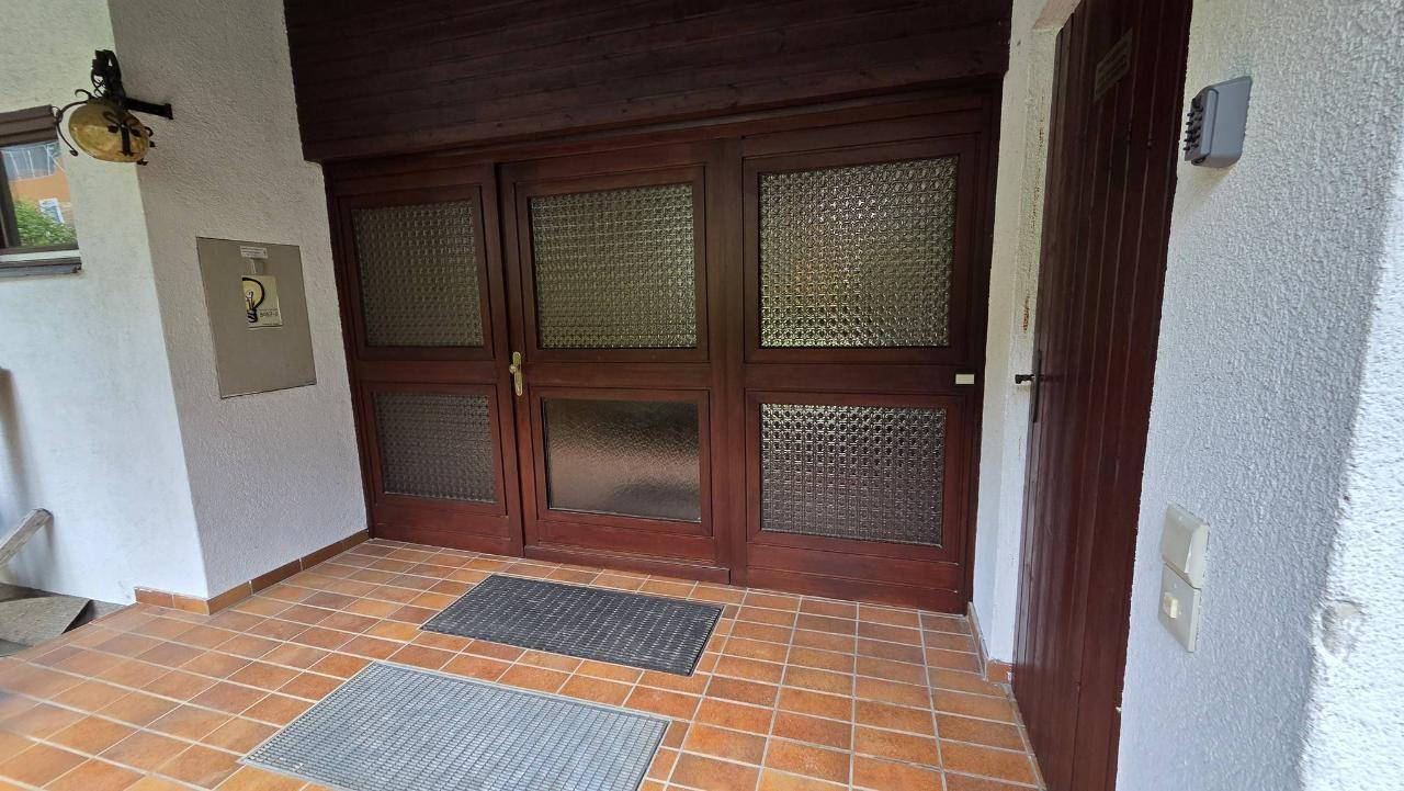 Eingangsbereich mit Holztüren, strukturiertem Glas und Fliesenboden, der zum Inneren führt.