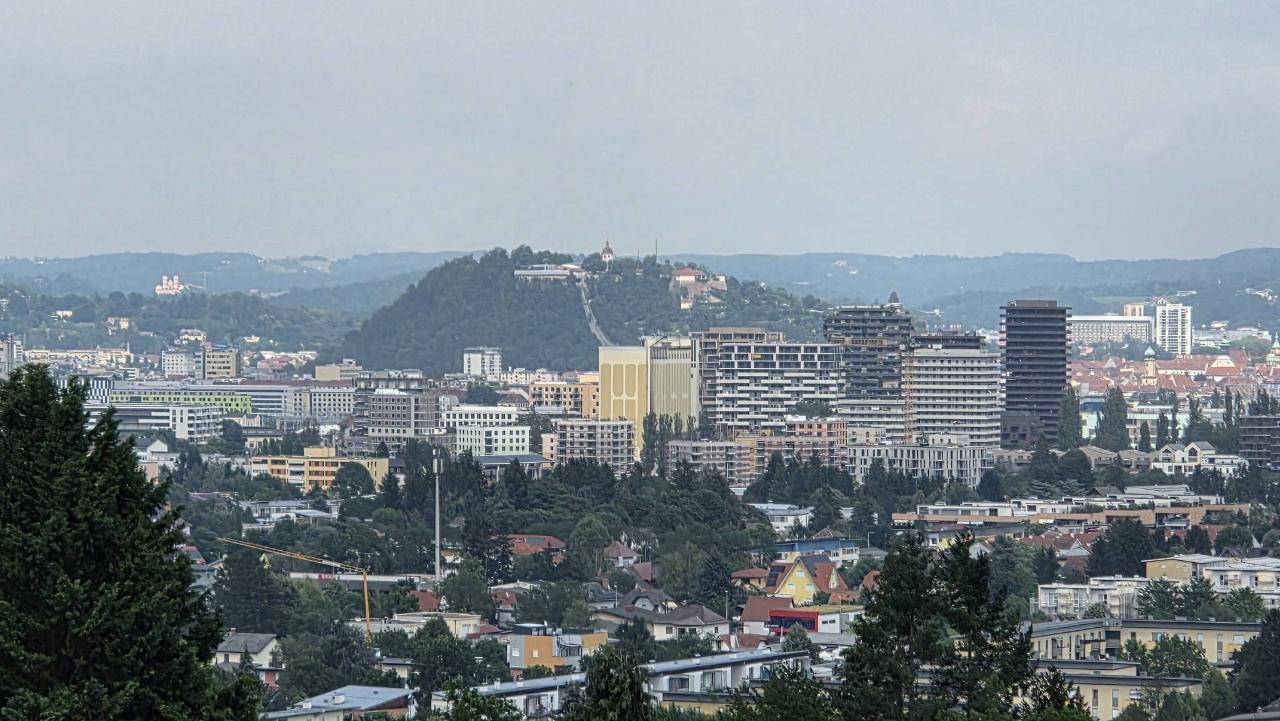 Panoramablick auf die städtische Skyline mit modernen Gebäuden und bewaldeten Hügeln im Hintergrund.