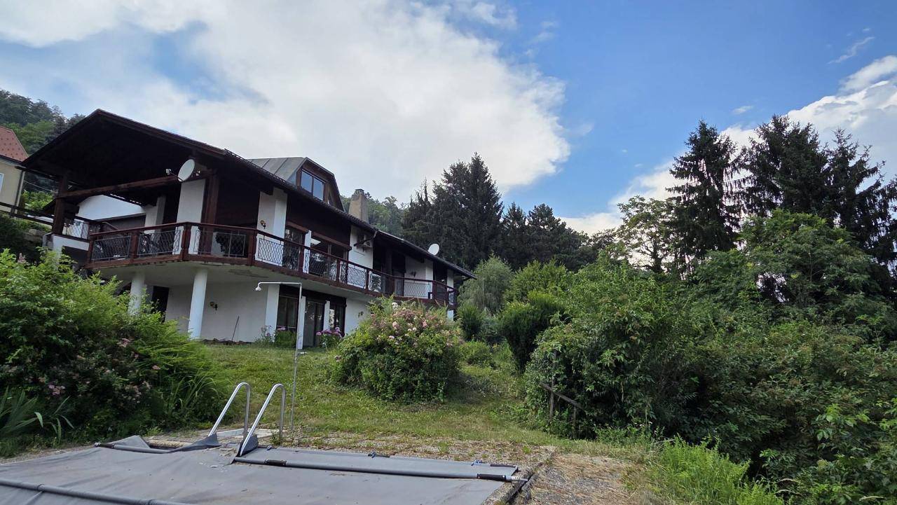 Hausfassade mit Balkonen und einem überdachten Pool im Gartenbereich unter blauem Himmel.