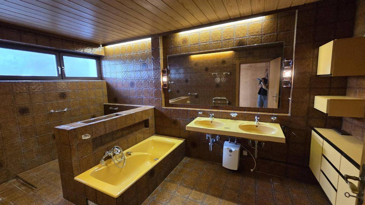 Badezimmer im 80er-Jahre-Stil mit braunen Fliesen, gelber Badewanne und Doppelwaschbecken.