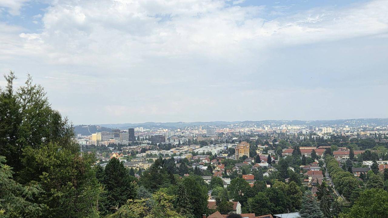 Panoramablick auf eine weitläufige Stadtlandschaft, umgeben von üppigem Grün und bewölktem Himmel.